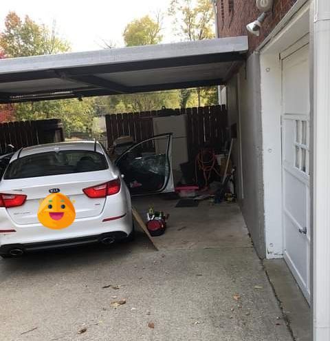 White sedan parked under a carport beside a garage, with tools and equipment cluttering the driveway.