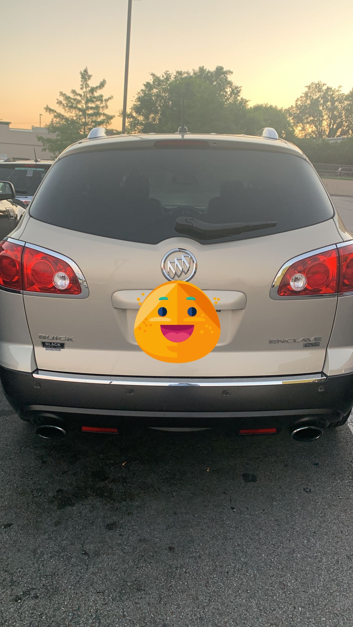 Silver SUV parked on a gravel lot at sunset, rear view with taillights and a smiley sticker on the license plate area