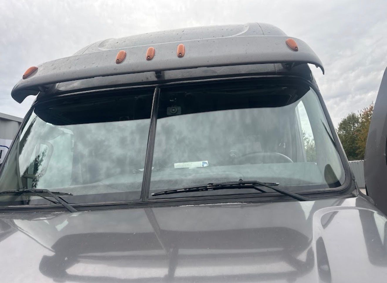 Front view of a gray truck cab with a roof visor and cloudy sky reflected in the windshield